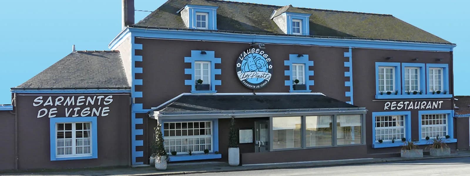 Façade de l'auberge Chez Pipette, bleue et brune, avec l'enseigne Sarments de vigne peinte au-dessus de la fenêtre