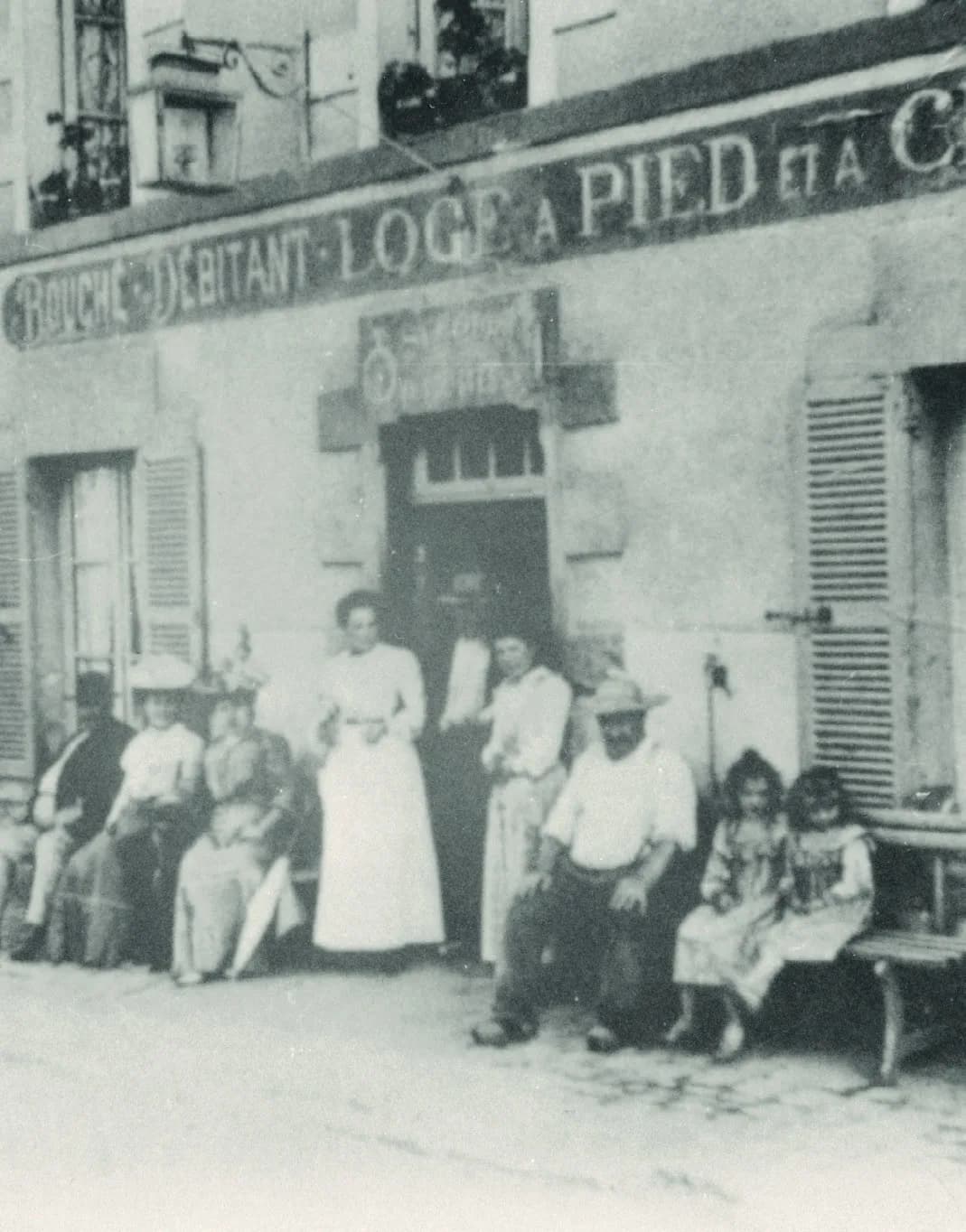 Photo ancienne en noir et blanc : la famille et des habitués assis devant l'auberge, sous l'enseigne On loge à pied et à cheval